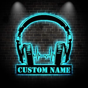 Puede incluir: Arte mural de metal negro que representa unos auriculares con un contorno de neón azul. Una línea de latidos del corazón está en el centro. El texto "CUSTOM NAME" se muestra en una base rectangular.