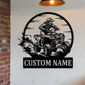 May include: Black metal wall art depicting an ATV rider in a circular frame with a sunburst design. The ATV is shown in motion, with water splashing. The text "CUSTOM NAME" is displayed on a rectangular plaque below the ATV.