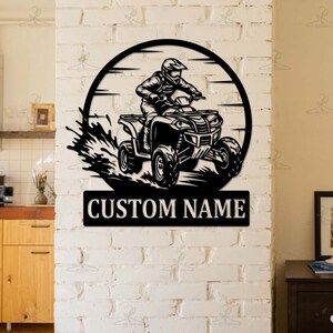 May include: Black metal wall art featuring an ATV rider within a circular frame. The design includes a splash effect and a rectangular plate with the text "CUSTOM NAME". The artwork is mounted on a white brick wall.