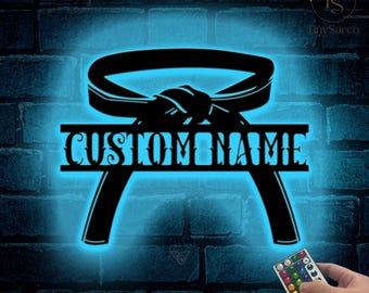 May include: Black silhouette of a martial arts belt with a custom name in a decorative font. The belt is tied in a knot and the name is centred below the belt. The image is illuminated with a blue light.
