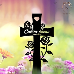 May include: Black metal cross with a heart cutout and rose accents. The cross features the text "Custom Name" and the years "2004-2024". The background is a soft blur of green, pink, and yellow.