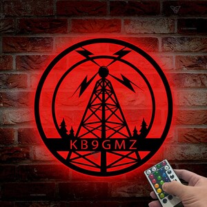 May include: A red illuminated round sign with a black silhouette of a radio tower and trees. The text "KB9GMZ" is displayed below the tower. A remote control with color changing LED lights is shown in the bottom right corner.