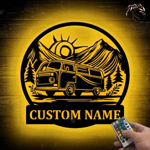 May include: Black and yellow metal wall art featuring a camper van, mountains, and sun design. The words "CUSTOM NAME" are displayed below the image. A remote control with colourful buttons is in the lower right corner.