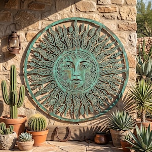 Puede incluir: Un gran adorno mural redondo de metal color verdín con un rostro solar y detalles intrincados de hojas y flores. El sol está montado en una pared de piedra texturizada, rodeado de cactus y suculentas en macetas.