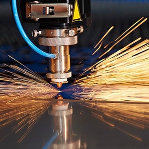 May include: A close-up of a laser cutter in operation. The machine is silver and blue, with a yellow warning label. Bright sparks emanate from the cutting point, reflecting on the metal surface below. The image captures the precision and power of industrial cutting.