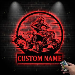 May include: A red, illuminated metal sign featuring a silhouette of a person riding an ATV within a circular frame. The sign includes the text "CUSTOM NAME" in a rectangular panel below the ATV image. The background is a dark brick wall.