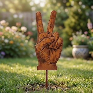 以下が含まれることがあります： 平和のサインをする手の形をした、さびた金属製のガーデンステーク。手には線やしわが細かく描かれ、風化した茶色の仕上がりです。ステークは緑の草に植えられ、背景にはぼやけた庭があります。