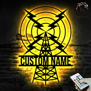 Op de afbeelding: Een zwart metalen silhouet van een radiotoren met bliksemschichten en concentrische cirkels, verlicht met geel licht. De tekst "CUSTOM NAME" wordt onder de toren weergegeven. Een afstandsbediening met kleurveranderende LED-verlichting is zichtbaar in de rechterbenedenhoek.