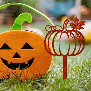 May include: An orange felt pumpkin-shaped container with a green handle and a carved jack-o'-lantern face. A rusty metal pumpkin stake with a leaf is in the grass, evoking a Halloween theme.