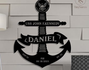 Op de afbeelding: Zwarte metalen anker met de tekst "USS JOHN F. KENNEDY" en "DANIEL" op een witte muur. Het anker heeft ook de tekst "SHIP 09", "PIR 08/18/2024" en "DIV 170".