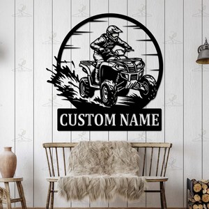 May include: Black metal wall art featuring an ATV rider within a circular frame. The ATV is depicted in motion, with a splash effect. Below the image is the text "CUSTOM NAME" in white letters, perfect for personalization.