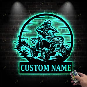 May include: Black metal ATV sign with a teal neon glow. The sign features a silhouette of a person riding an ATV, with the words "CUSTOM NAME" below. The sign is set against a brick wall background.