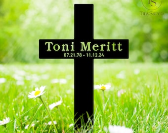 以下が含まれることがあります： 白いプレートに「Toni Merritt」という名前と「07.21.78 - 11.12.24」という日付が刻まれた黒い金属製の十字架。十字架は、白い花が咲く草地の中に置かれている。