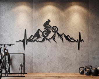 May include: A black metal wall art piece featuring a silhouette of a mountain biker riding over a mountain range, integrated into a continuous heartbeat EKG line design. The artwork is mounted on a gray concrete wall above a wooden floor.