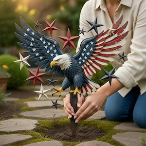 Puede incluir: Una estaca de jardín patriótica con un águila calva detallada con las alas extendidas, pintada en rojo, blanco y azul. Las alas muestran estrellas y rayas, y el águila está rodeada de adornos en forma de estrella. La estaca se está colocando en el suelo.