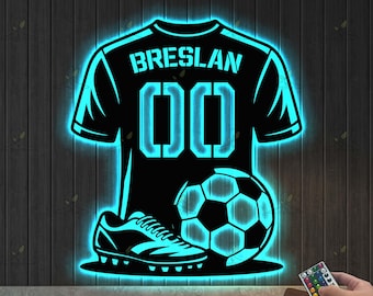 May include: Black soccer-themed wall art with a neon blue outline. The design features a soccer jersey with the name "Breslan" and the number "00", a soccer ball, and a soccer cleat. A remote control is visible in the bottom right corner.