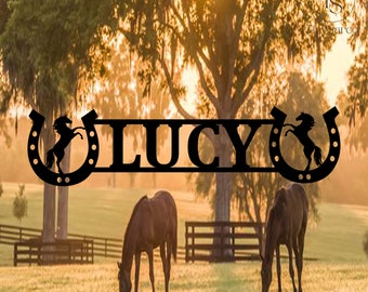 Pode incluir: Um letreiro de metal preto com o nome "LUCY" em letras grandes, ladeado por duas ferraduras com uma silhueta de um cavalo dentro de cada ferradura.