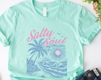 May include: A light green t-shirt featuring a graphic design of a palm tree, sun, and waves. The text "Salty Soul" is printed in pink on the shirt.