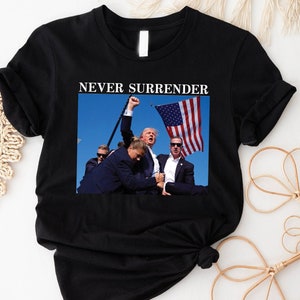May include: Black t-shirt with a white print that says "NEVER SURRENDER". The print also includes a photo of a man holding an American flag.