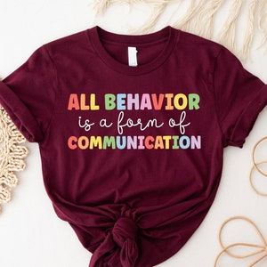 May include: A maroon t-shirt with the text "ALL BEHAVIOR is a form of COMMUNICATION" in rainbow colors.