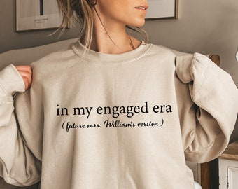 Könnte beinhalten: Ein beiger Pullover mit dem Aufdruck "in my engaged era (future mrs. William's version)" auf der Vorderseite.