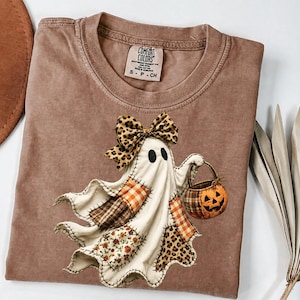 May include: A brown t-shirt featuring a graphic of a ghost wearing a leopard print bow and holding a jack-o'-lantern bucket. The ghost is decorated with various fabric patches including plaid, floral, and leopard print patterns. The shirt has a Comfort Colors label.