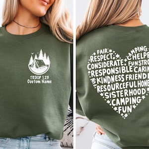 May include: Olive green sweatshirt with a white graphic of a tent and trees, with the text "TROOP 123 Custom Name". The back of the sweatshirt features a heart-shaped design with words like "Camping", "Kindness", and "Fun".