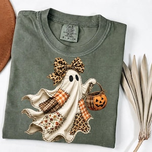 May include: An olive green t-shirt featuring a graphic of a white ghost wearing a leopard print bow and carrying a pumpkin bucket. The ghost has patchwork sections in plaid, floral, and leopard patterns. The shirt has a Comfort Colors brand label.