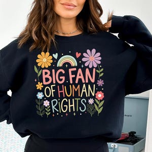 May include: Black sweatshirt with the text "BIG FAN OF HUMAN RIGHTS" in colorful letters. The design includes flowers, a rainbow, and hearts. The sweatshirt is a comfortable, casual style.