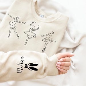 May include: A beige sweatshirt with three black line drawings of ballerinas and the word "Melissa" printed on the sleeve. The sleeve also has a black line drawing of a pair of ballet shoes.