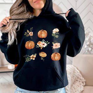 Puede incluir: Sudadera con capucha negra con un dise&ntilde;o de calabazas y arreglos florales en tonos naranja, melocot&oacute;n y verde. El dise&ntilde;o est&aacute; impreso en la parte delantera. La sudadera tiene un bolsillo delantero y una capucha con cord&oacute;n.