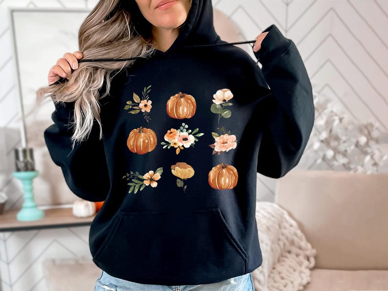 Puede incluir: Sudadera con capucha negra con un dise&ntilde;o de calabazas y arreglos florales en tonos naranja, melocot&oacute;n y verde. El dise&ntilde;o est&aacute; impreso en la parte delantera. La sudadera tiene un bolsillo delantero y una capucha con cord&oacute;n.
