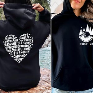 May include: Black hoodies with white graphics. One features a heart-shaped design with words like "Camping" and "Kindness." The other displays a tent and trees with "TROOP 123" below. Both have a front pocket and hood.