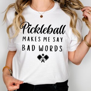 May include: White t-shirt with black text that reads "Pickleball makes me say bad words" and a graphic of two pickleball paddles and a ball.