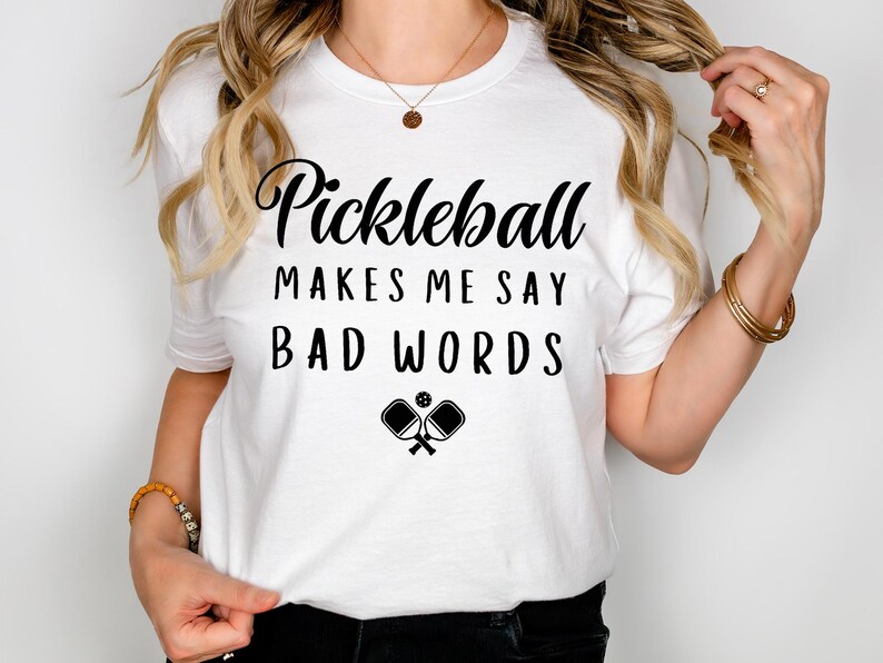 May include: White t-shirt with black text that reads "Pickleball makes me say bad words" and a graphic of two pickleball paddles and a ball.
