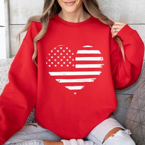 May include: Red sweatshirt with a white distressed heart-shaped American flag design.