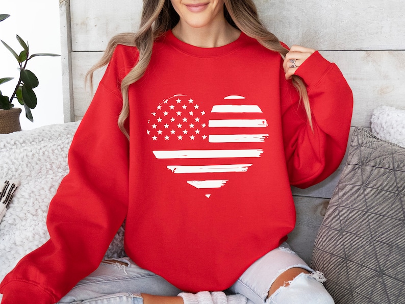 May include: Red sweatshirt with a white distressed heart-shaped American flag design.