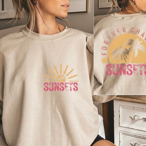 May include: A beige sweatshirt with a graphic print of a sun and palm trees. The text "Forever Chasing Sunsets" is printed on the back of the sweatshirt. The text "Sunsets" is printed on the front of the sweatshirt.