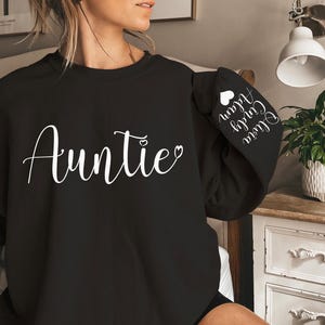 May include: Black sweatshirt with the word "Auntie" in white cursive lettering and a small heart. The left sleeve has the text "Our Girl" and "Auntie" in white cursive lettering with a small heart.