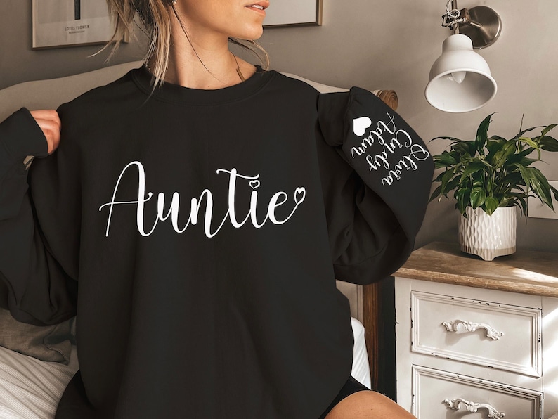 May include: Black sweatshirt with the word "Auntie" in white cursive lettering and a small heart. The left sleeve has the text "Our Girl" and "Auntie" in white cursive lettering with a small heart.
