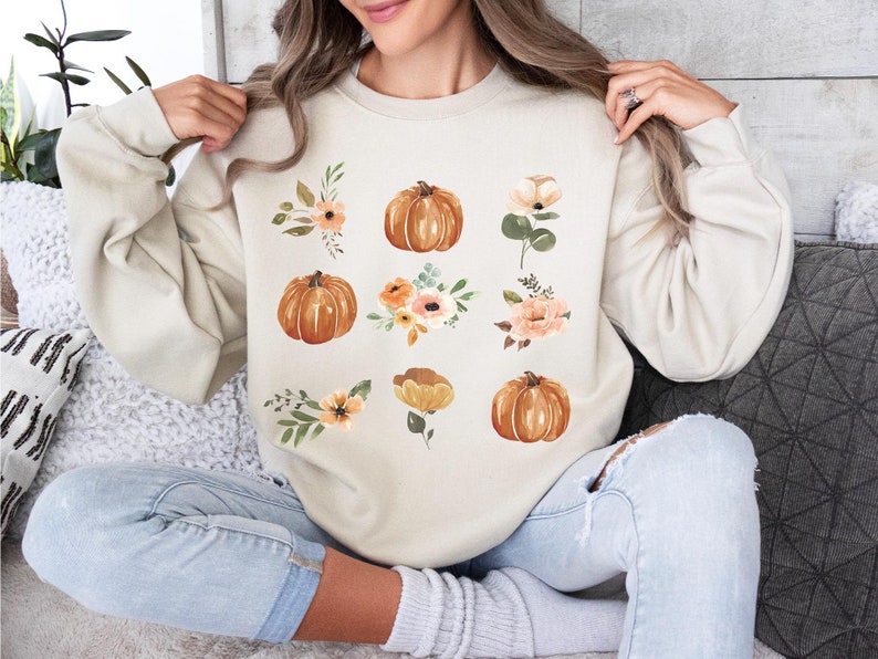 Puede incluir: Sudadera beige con un dise&ntilde;o de tem&aacute;tica oto&ntilde;al. El dise&ntilde;o incluye ilustraciones de acuarela de calabazas en tonos naranja y arreglos florales en melocot&oacute;n, naranja y verde. La sudadera es de corte holgado.