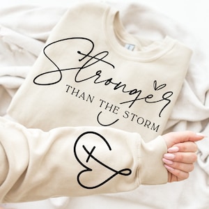 May include: A beige sweatshirt with the text "Stronger than the storm" and a heart with a cross inside on the sleeve.