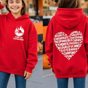 May include: Red hooded sweatshirt with a front pocket and white graphic. The front graphic includes a tent and trees with the text "TROOP 123 Custom Name". The back features a heart-shaped design with words like "Camping" and "Fun".