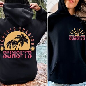 May include: Black hoodie with a graphic print of palm trees and a sunset on the back. The front of the hoodie has a graphic print of a sunset with palm trees and the text "SUNSETS".