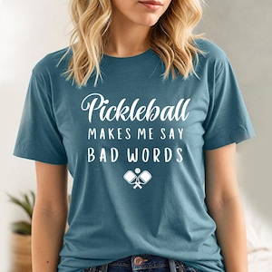 May include: A teal blue t-shirt with the text "Pickleball makes me say bad words" in white lettering. The shirt also features a white graphic of two pickleball paddles and a ball.