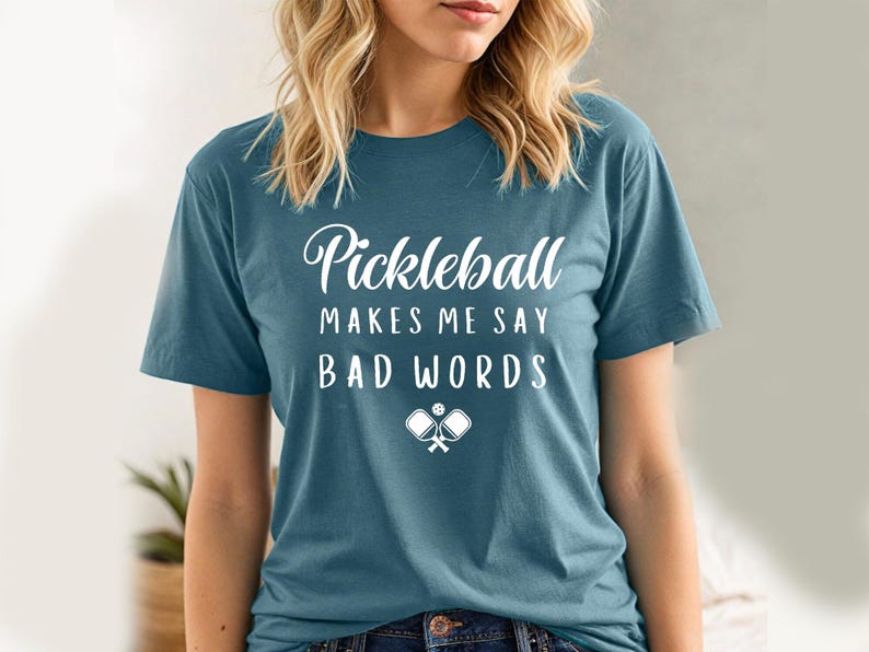 May include: A teal blue t-shirt with the text "Pickleball makes me say bad words" in white lettering. The shirt also features a white graphic of two pickleball paddles and a ball.