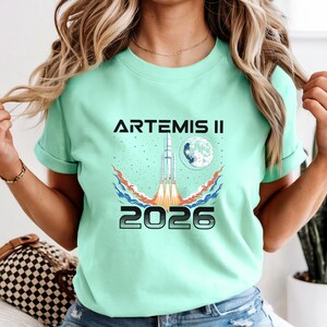 May include: A mint green t-shirt with the text "ARTEMIS II 2026" in black and silver. The graphic features a rocket launching towards a moon, with red, orange, and blue flames. The shirt is a crew neck style.
