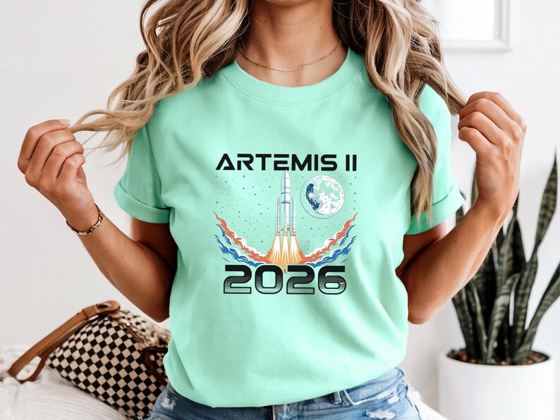 May include: A mint green t-shirt with the text "ARTEMIS II 2026" in black and silver. The graphic features a rocket launching towards a moon, with red, orange, and blue flames. The shirt is a crew neck style.