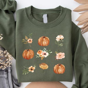 Puede incluir: Una sudadera verde oliva con un dise&ntilde;o de acuarela de calabazas y detalles florales. Las calabazas son de color naranja y las flores son de color melocot&oacute;n y blanco. El dise&ntilde;o se repite en la parte delantera de la sudadera. Prenda casual.
