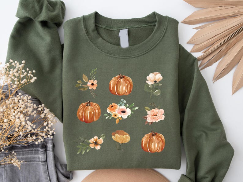 Puede incluir: Una sudadera verde oliva con un dise&ntilde;o de acuarela de calabazas y detalles florales. Las calabazas son de color naranja y las flores son de color melocot&oacute;n y blanco. El dise&ntilde;o se repite en la parte delantera de la sudadera. Prenda casual.